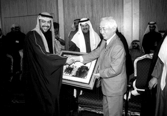 新契約調印式ののち、記念品を交換する小長社長とシエイク・アハマド・アルサバーハ石油大臣代行の画像が表示されています。